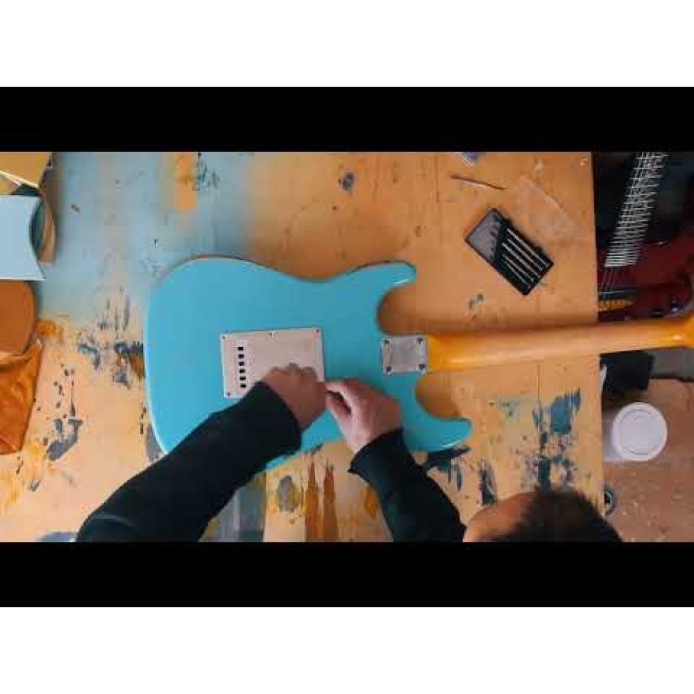 Hands installing white pickguard onto turquoise guitar body during assembly build process.